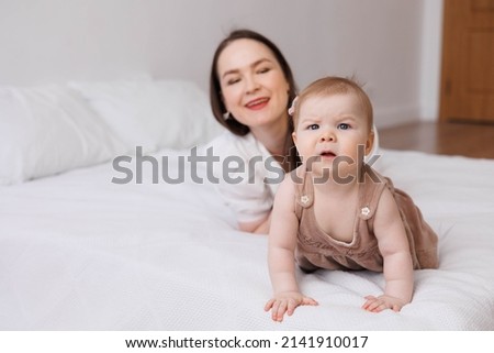 Sweet young mother playing with her little 1-year-old daughter in bed.
Caring brunette woman laying with her baby child at home. Tenderness and love between mother and daughter. Lovely family indoors.