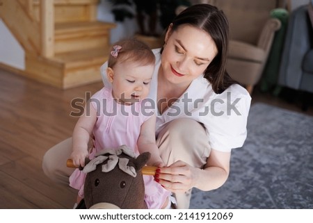 Sweet young mother playing with her little 1-year-old daughter.
Caring brunette woman help her baby child to ride a toy horse. Tenderness and love between mother and daughter. Lovely family indoors.