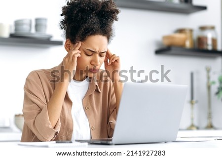 Tired african american girl overworked from working in a laptop, sitting at a table in the kitchen at home, closed her eyes, massages her temples, has a migraine, headache, tension, needs rest, break