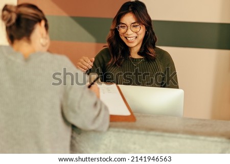 Cheerful receptionist assisting a woman with signing in to an office. Friendly receptionist showing a woman where to sign on a clipboard. Young woman working at the front desk of a co-working space.