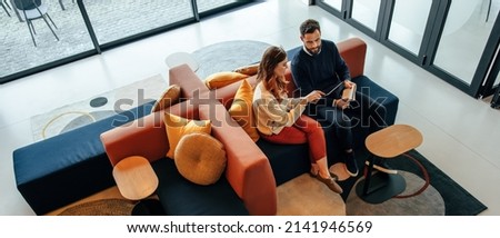 High angle view of two businesspeople working in an office lobby. Two modern businesspeople using a digital tablet while having a discussion. Two young entrepreneurs collaborating on a new project. Royalty-Free Stock Photo #2141946569