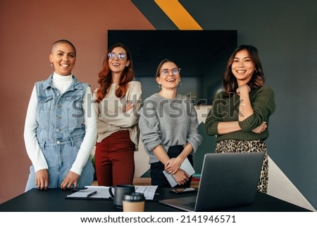 Successful female entrepreneurs smiling cheerfully while standing behind a table in a boardroom. Group of multicultural businesswomen working together in a modern office. Royalty-Free Stock Photo #2141946571