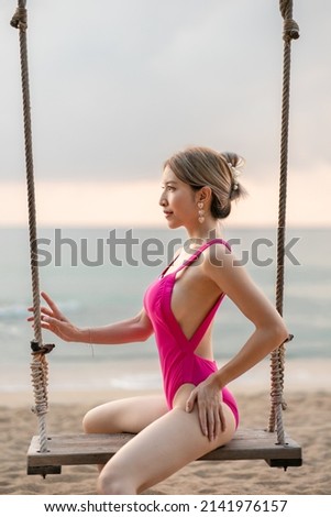 Woman in pink one piece swimsuit enjoying on swing on the beach, tropical island. Over the ocean background.