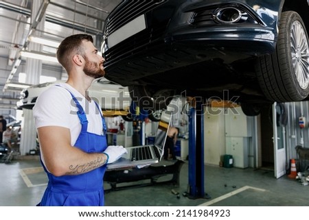 Happy technician mechanic man 20s wear blue overalls t-shirt use hold laptop pc computer stand near car lift check technical condition work in vehicle repair shop workshop indoors Tattoo translate fun Royalty-Free Stock Photo #2141984923