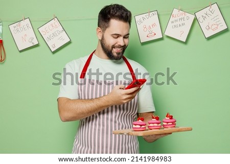 Young happy chef confectioner baker man in apron hold three cake muffin macaroon on board taking photo picture on mobile cell phone isolated on plain pastel light green background Cooking food concept