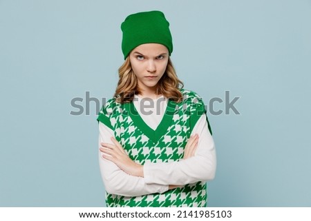 Angry unnerved aggrieved disconcerted sad young brunette girl teen student wears checkered green vest hat look camera hold hands crossed isolated on plain pastel light blue background studio portrait Royalty-Free Stock Photo #2141985103