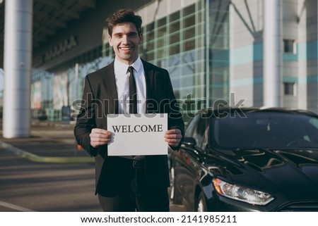 Bottom view young smiling friendly traveler businessman man 20s in black suit stand outside at international airport terminal hold card sign with welcome title text Air flight business trip concept.