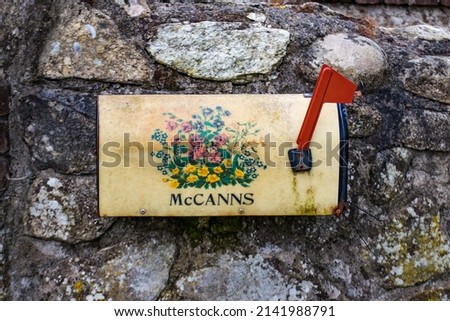 Old weathered metal mailbox mounted on gray stone wall with red flag and flowers paint motive. Ireland