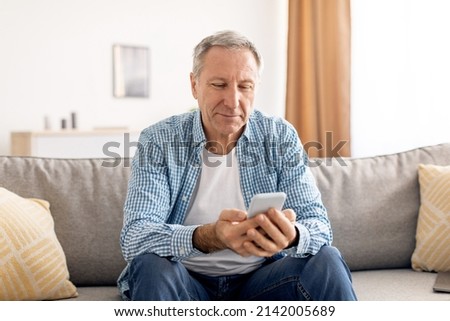 People And Technology. Portrait of smiling mature man using mobile phone, watching video or reading sms message, sitting on the couch in living room at home. Adult guy browsing internet, surfing web