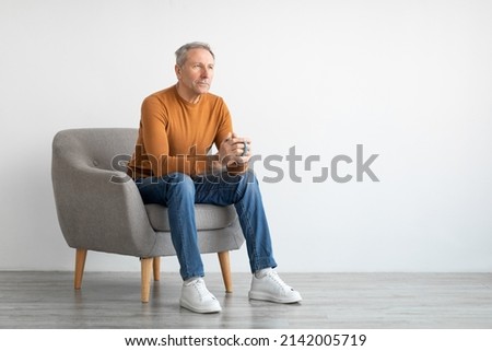 Mature man drinking coffee sitting on chair in living room. Casual adult guy relaxing, enjoying weekend free time, mockup free copy space, full body length, isolated on white studio background wall