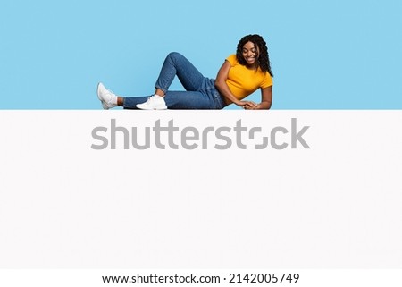 Relaxed pretty millennial african american woman with curly hair chilling on empty white horizontal advertising board, looking down at copy space and smiling, mockup, blue studio background
