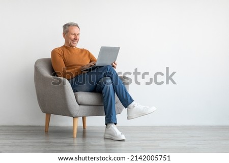 Smiling mature man using laptop sitting on armchair in living room. Happy confident senior male adult resting at home working on pc, typing on keyboard isolated on white studio wall, full body length Royalty-Free Stock Photo #2142005751