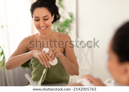 Portrait of smiling young woman applying moisturizing cream or lotion on her hands near mirror at home, free space. Charming millennial lady taking care of her skin with nourishing creme at bathroom