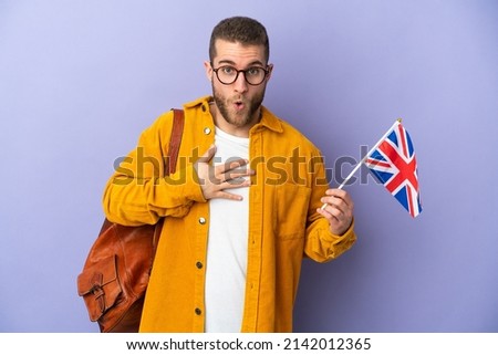 Young caucasian man holding an United Kingdom flag isolated on purple background surprised and shocked while looking right
