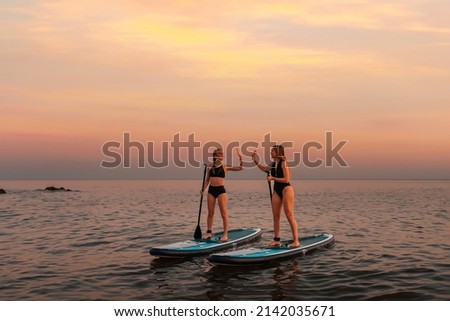 Two happy women stands on a sup boards and high five with hands. In the background, the ocean and the sunset. Copy space. Summer extreme recreation and surfing. Royalty-Free Stock Photo #2142035671