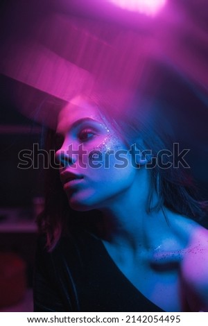 Fashionable photo of a woman with bright makeup in neon light with purple and blue in a dark room, looking away. vertical