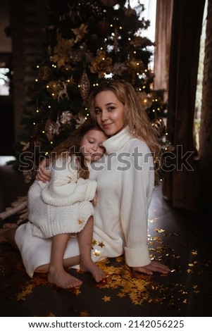 Cute mom and daughter in bright knitted clothes playing with golden confetti stars on the background of a decorated Christmas tree. noise effect, selected focus.