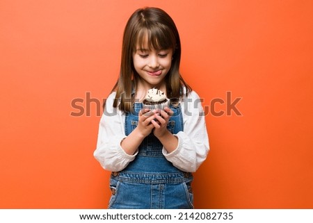 Adorable little girl with her tongue out and looking at a delicious chocolate cupcake with a smile Royalty-Free Stock Photo #2142082735