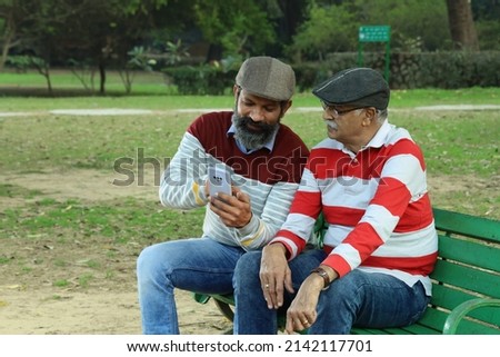 Father and mid aged son having a good time sitting on a park bench holding a mobile phone in hand. they are surrounded with lush greens and serene environment.