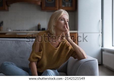Frustrated lonely mature 50s woman looking away, sitting on sofa at home touching face. Sad lady thinking over health problems, suffering from apathy, depress disorder, memory loss, dementia Royalty-Free Stock Photo #2142121549