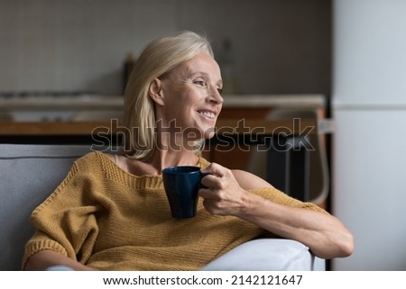 Happy dreamy senior blonde lady enjoying good morning, drinking hot coffee at home, resting on comfortable couch in cozy living room, holding ceramic mug, cup of tea, smiling, looking away