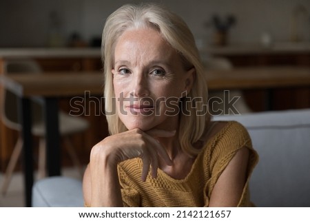 Smiling blonde middle aged 60s lady home head shot portrait. Skinny retired woman, pensioner, OAP sitting on couch, touching chin, looking at camera, posing for profile picture Royalty-Free Stock Photo #2142121657