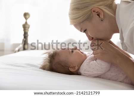 Happy pretty young mom cuddling sweet baby lying on back on bed, holding, kissing infant arms. Devoted mum playing with little child with love, affection, joy, enjoying motherhood, family time