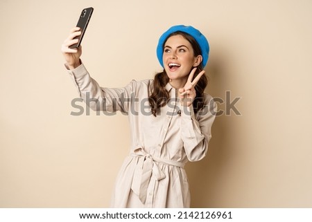 Beautiful modern woman posing for selfie, holding smartphone, taking picture photo on mobile phone, standing happy against beige background