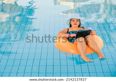brunette jewish woman in a sun hat in the pool in the swimming circle with a cocktail and a laptop relaxes. Horizontal photo.