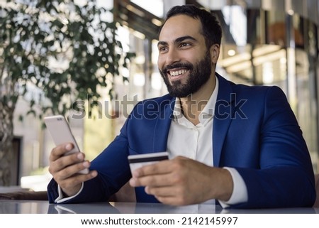 Handsome smiling Arabian man holding credit card using mobile phone shopping online check card balance. Mobile banking. Successful middle eastern freelancer receive payment sitting at workplace