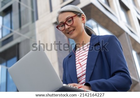 Smiling confident asian businesswoman using laptop computer working online on the street. Mature grey haired woman wearing stylish eyeglasses having video call sitting at workplace 