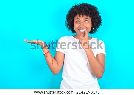 Positive young woman with afro hairstyle wearing white T-shirt against blue wall advert promo touches teeth with finger.