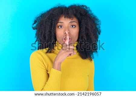 Young woman with afro hairstyle wearing yellow turtleneck over blue background makes silence gesture, keeps finger over lips. Silence and secret concept.