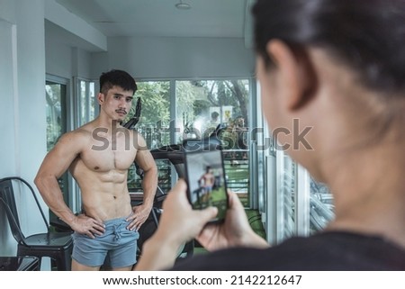 A woman taking a picture of a fitness influencer flaunting his impressive physique with her camera, to be posted on social media. A scene at the gym.