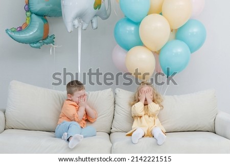 Blonde girl and a boy hide their faces in hands on a sofa in a white studio with yellow and green balloons. Festive decoration. Children's happiness. Valentine's Day.