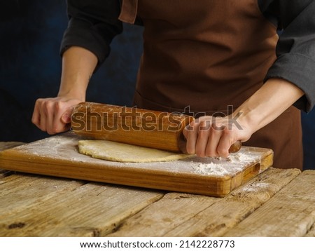 The chef in a dark uniform rolls out the dough with a rolling pin on a cutting board, wooden background. Preparation of dough products - pizza, pie, ravioli, confectionery.