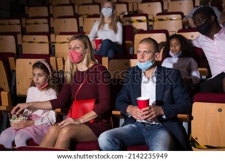Friendly family in protective face masks eating popcorn and watching movie in cinema. Coronavirus pandemic safety rules and social distance concept