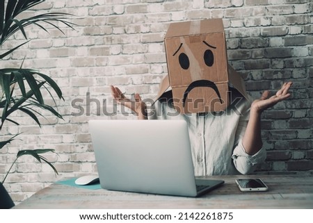 Modern worker with carton sad box on head open arms with desolation gesture in front of a laptop. Online crypto smart working business job activity. Concept of fail and lost work. Bad day office Royalty-Free Stock Photo #2142261875