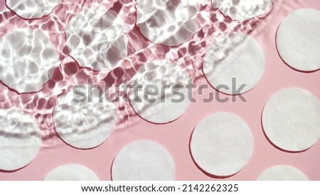 Wave flowing on water surface from left upper corner and making ripples over cotton pads arranged in rows on pink background | Background for beauty care product