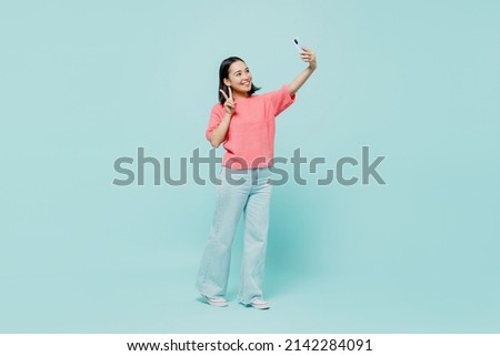 Full body young smiling happy woman of Asian ethnicity 20s wearing pink sweater doing selfie shot on mobile cell phone post photo on social network isolated on pastel plain light blue color background