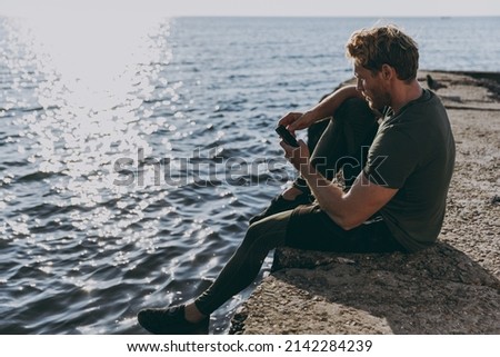 Full size side view young strong sporty fit sportsman man in sports clothes warm up training sit on pier use mobile cell phone at sunrise sun dawn over sea beach outdoor seaside in summer day morning.