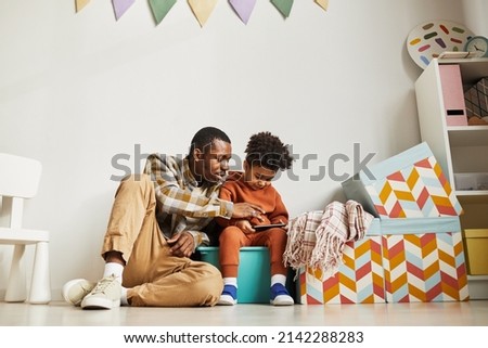 Full length portrait of African American father with little son using smartphone in cute kids room interior, copy space
