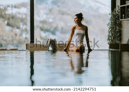 Attractive young woman in bikini sits by the poolside in the indoor swimming pool at winter time