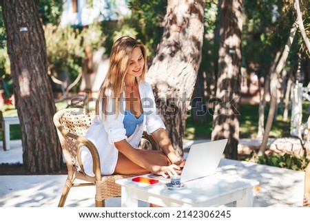 Beautiful smiling woman freelancer working on a laptop, typing on a keyboard, sitting in the outdoor cafe. Remote work and work from home concept.