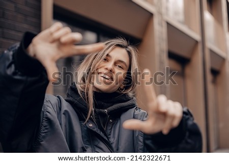 Attractive woman with a lovely smile making a frame gesture with her fingers framing her face. Woman posing on selfie photo looking at camera walking outdoors in urban city.