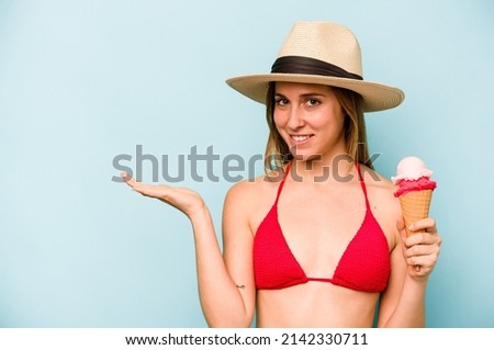 Young caucasian woman wearing a bikini and holding an ice cream isolated on blue background showing a copy space on a palm and holding another hand on waist.