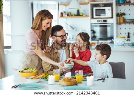 Family of four at the table, mother serves them breakfast. Everyone is happy and smiling, parents enjoy spending time with their children. A cheerful family has fun during a meal at the dining table. Royalty-Free Stock Photo #2142343401