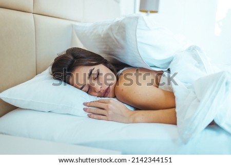 Horizontal shot of relaxed carefree female under white bedclothes in bed at bedroom, sees pleasant dreams, keeps eyes closed, enjoys good rest at home and calm atmosphere. People and rest concept