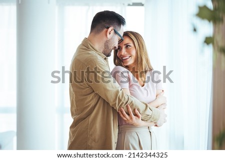 A pair of lovers, hug, kiss, laugh and have fun. Light gray interior, large window, brown stylish chair. Happy young couple standing near window at home