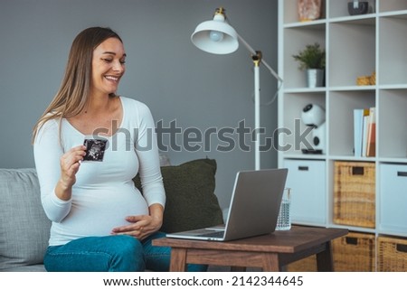 A young beautiful pregnant woman using a laptop on a gray sofa with an ultrasound image in a side view with copy space. Pregnancy, love, health care, people, the concept of relaxation.